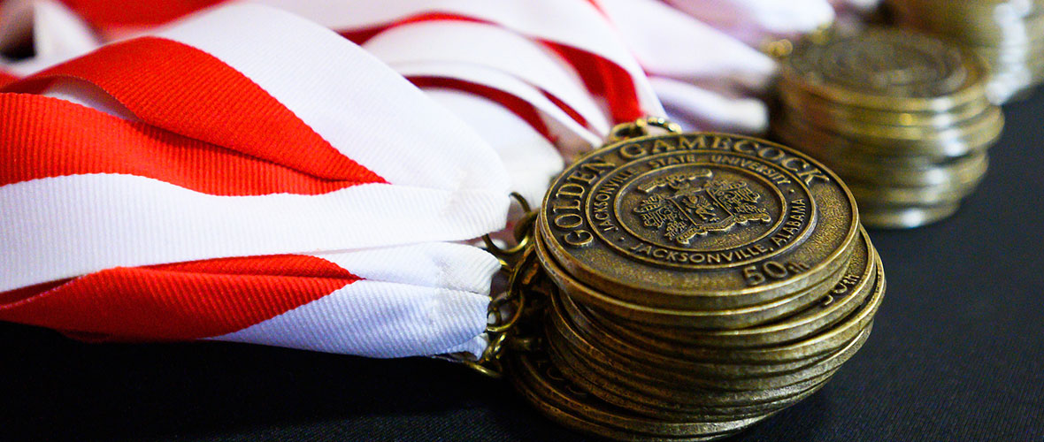 Golden Gamecock medals on bright red and white ribbons lie stacked on a table.