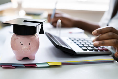 A piggy bank, wearing a graduation cap, along with a student on a laptop