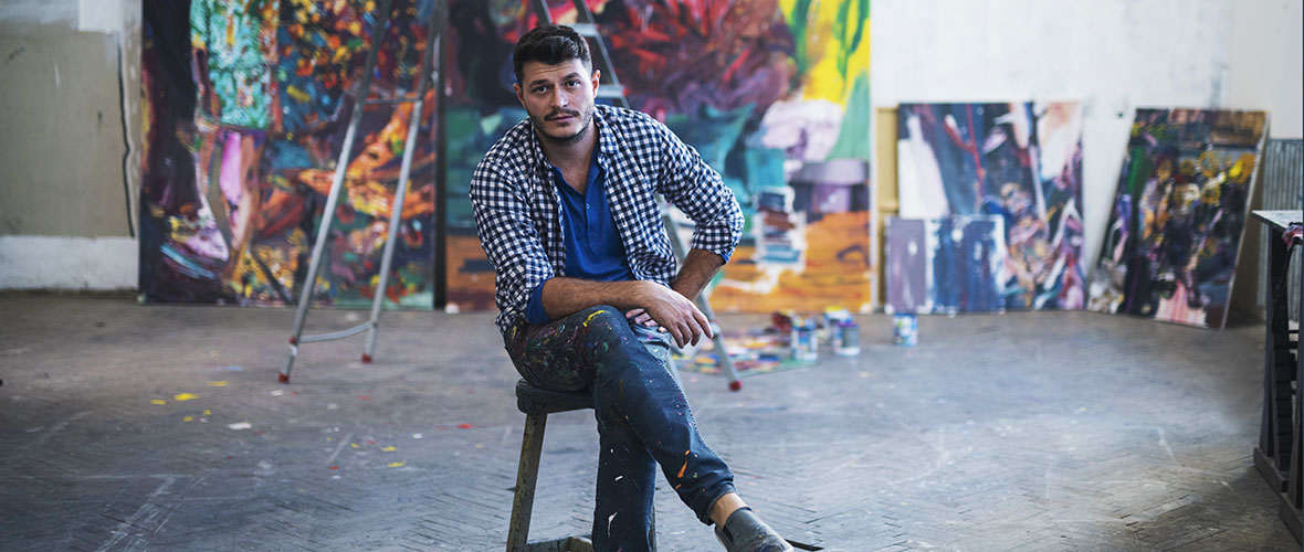 A male painter wearing paint-splattered jeans, perches on a stool in his studio, in front of his large works of art. 