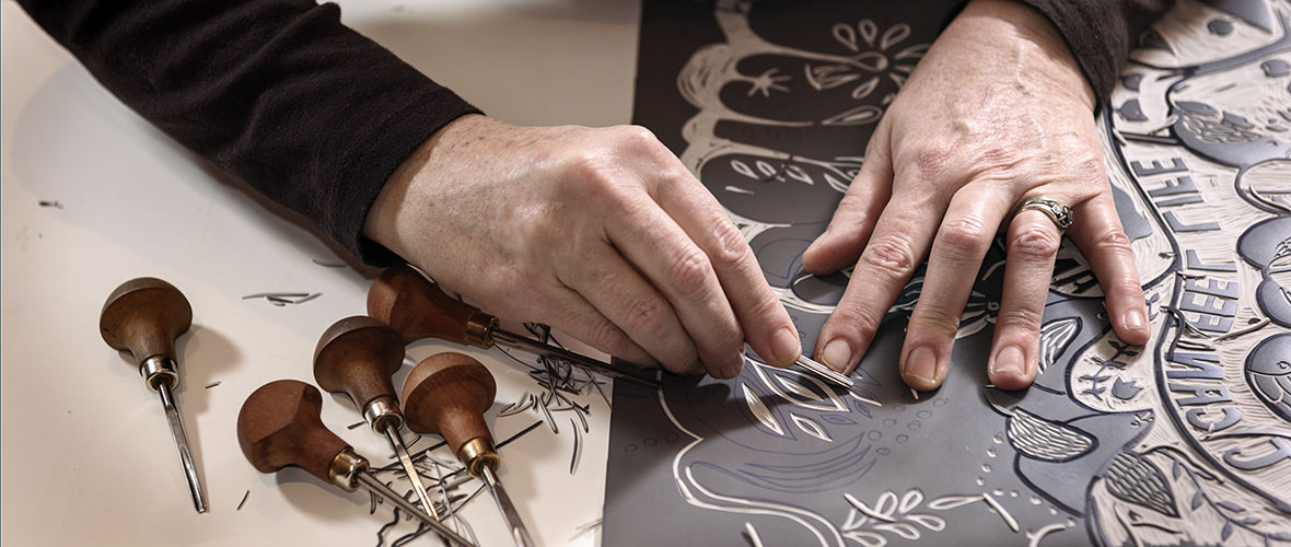 A close up of a printmaker artist's hands carving intricate animal and floral designs into a piece of linoleum