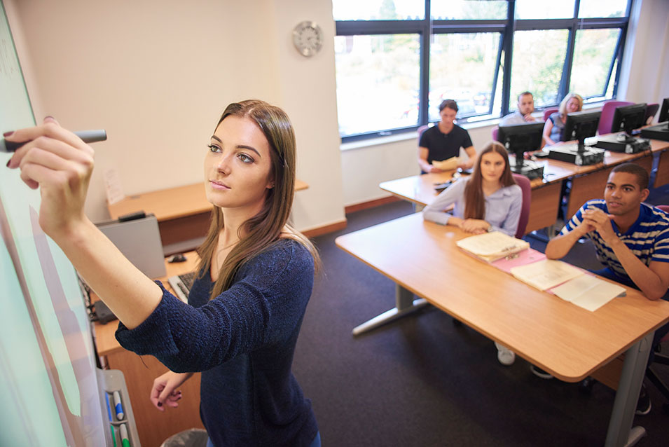 A student teacher writes on the board, her class looking on