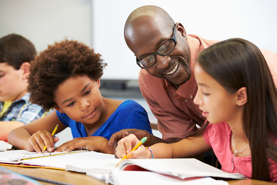 A male teacher assists two young students at their desks