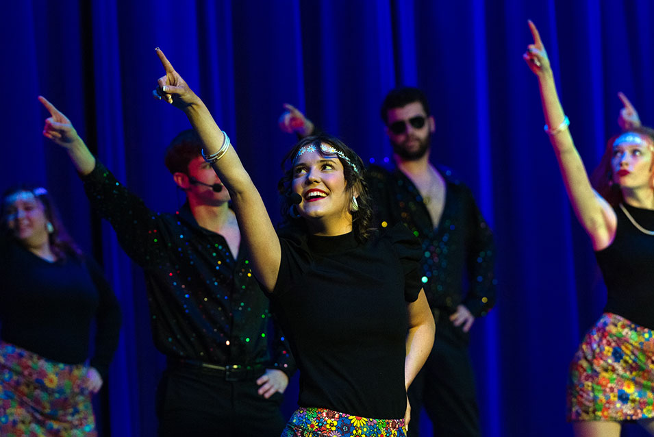 Female show choir members perform under blacklight wearing colorful feather boas around their necks