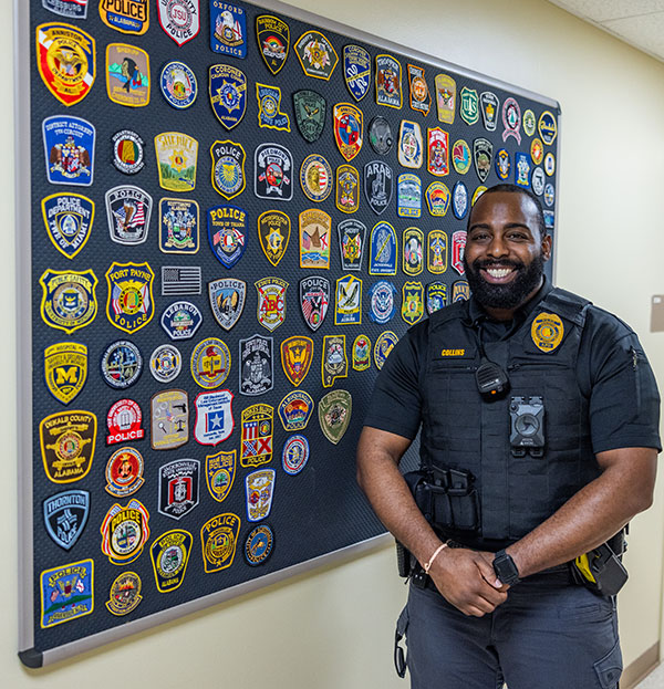 Anniston Police Department officer Collins poses in front of the wall of patches representing all of the departments served by LECC.