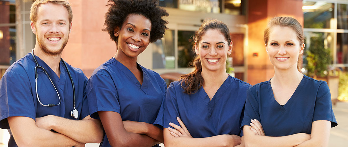 Three female nurses and a male nurse wearing a stethoscope stand outside an emergency room entrance.
