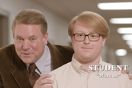 An older man stands behind a student in a hallway