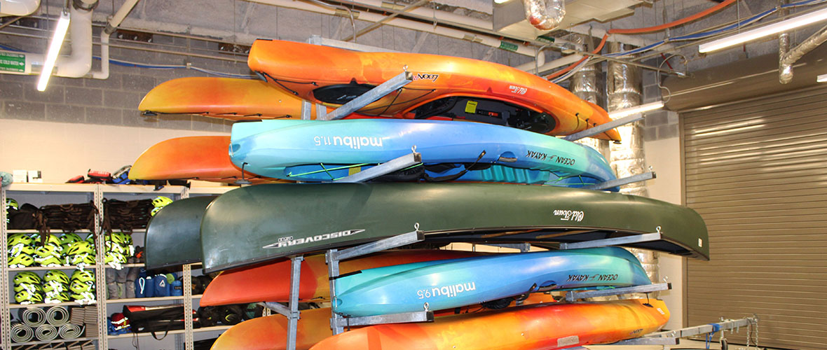 orange, blue, and green kayaks and canoes loaded on boat trailer inside a camping gear storage room.