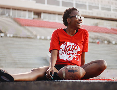 A female JSU student wearing a Jax State tee sits on the field at JSU stadium with a football in front of her